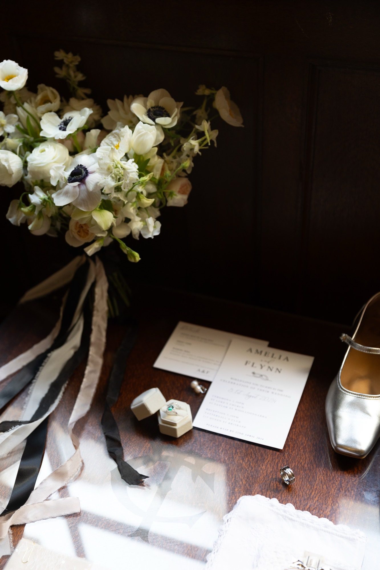Bridal bouquet of white anemones and ranunculus with black-and-white silk ribbons, beside the invitation suite, emerald engagement ring and silver shoes — Amelia and Flynn's wedding, Brisbane