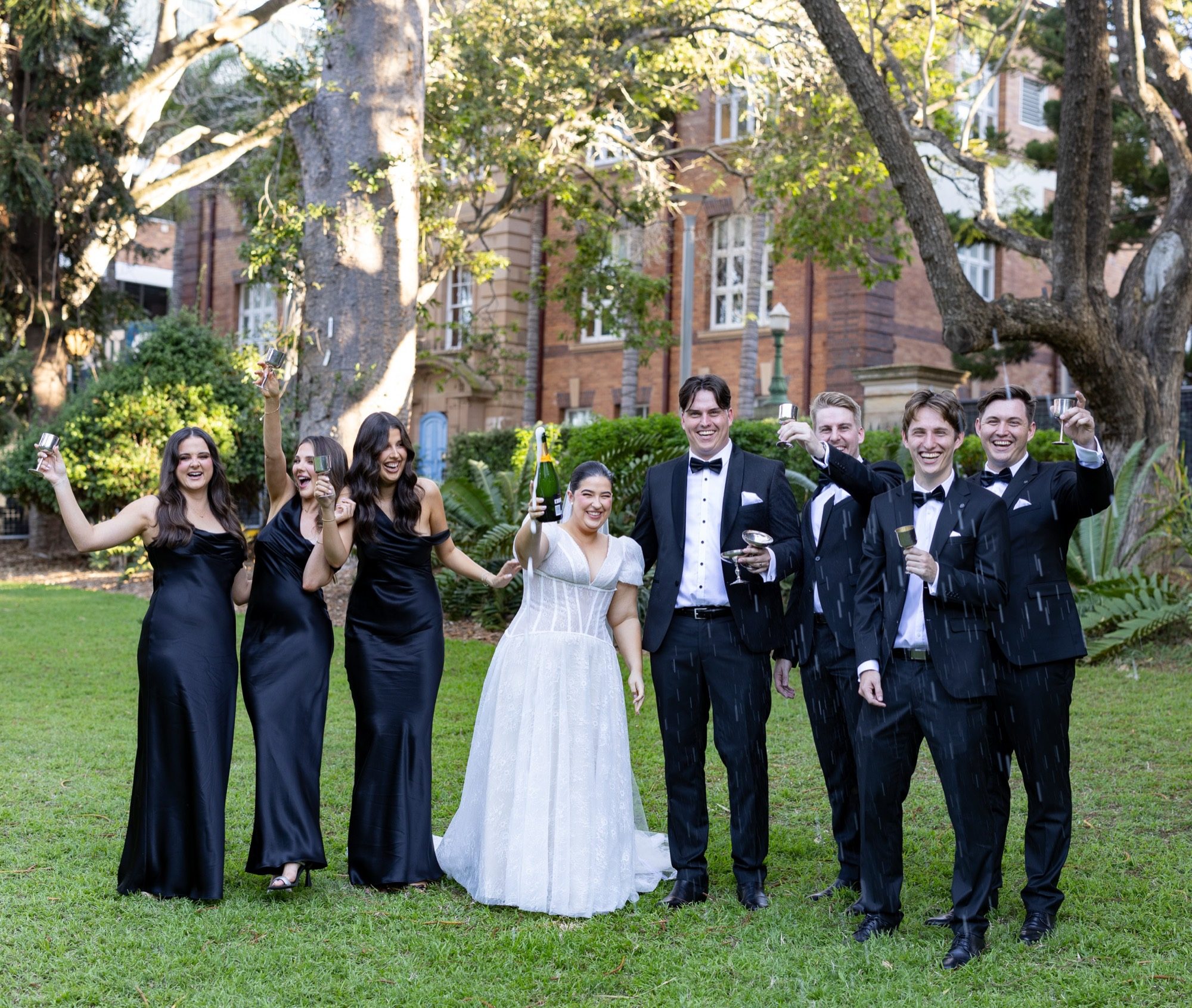The bridal party raising silver coupes as champagne is sprayed, beneath the fig trees outside a heritage brick building, Brisbane