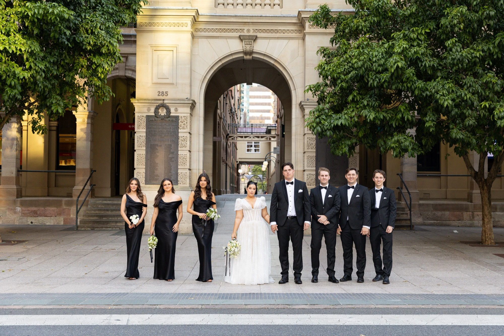 The full bridal party in black tie beneath the sandstone arch on Queen Street, Brisbane