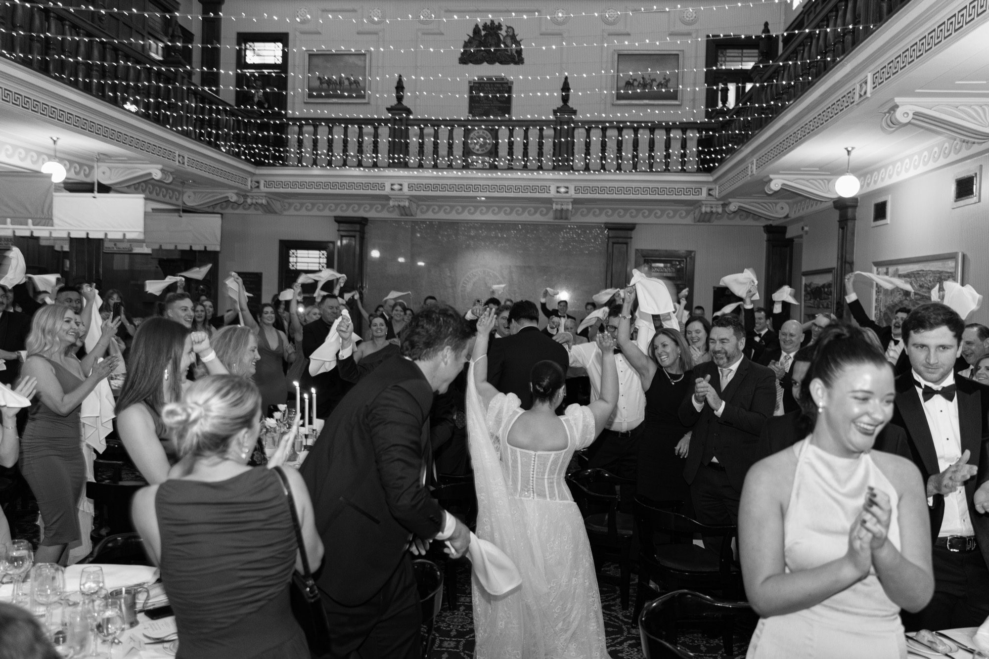 Guests waving white napkins as the couple enter the reception beneath the fairy-lit balcony, Tattersall's Club, Brisbane