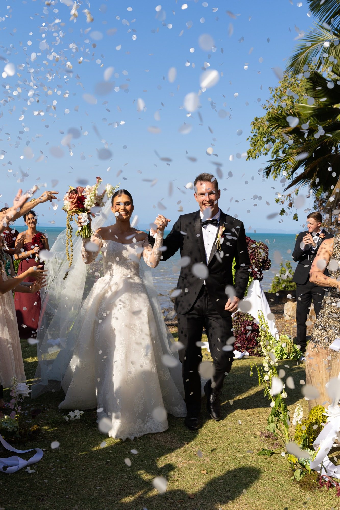 Confetti exit — the couple hand in hand after the ceremony with the sea behind them, Freedom Shores, Airlie Beach