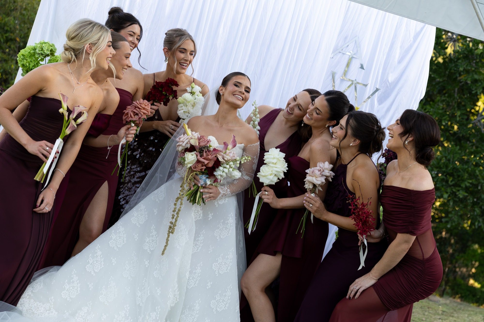 The bride laughing with her bridesmaids in burgundy before the draped monogram backdrop, Airlie Beach